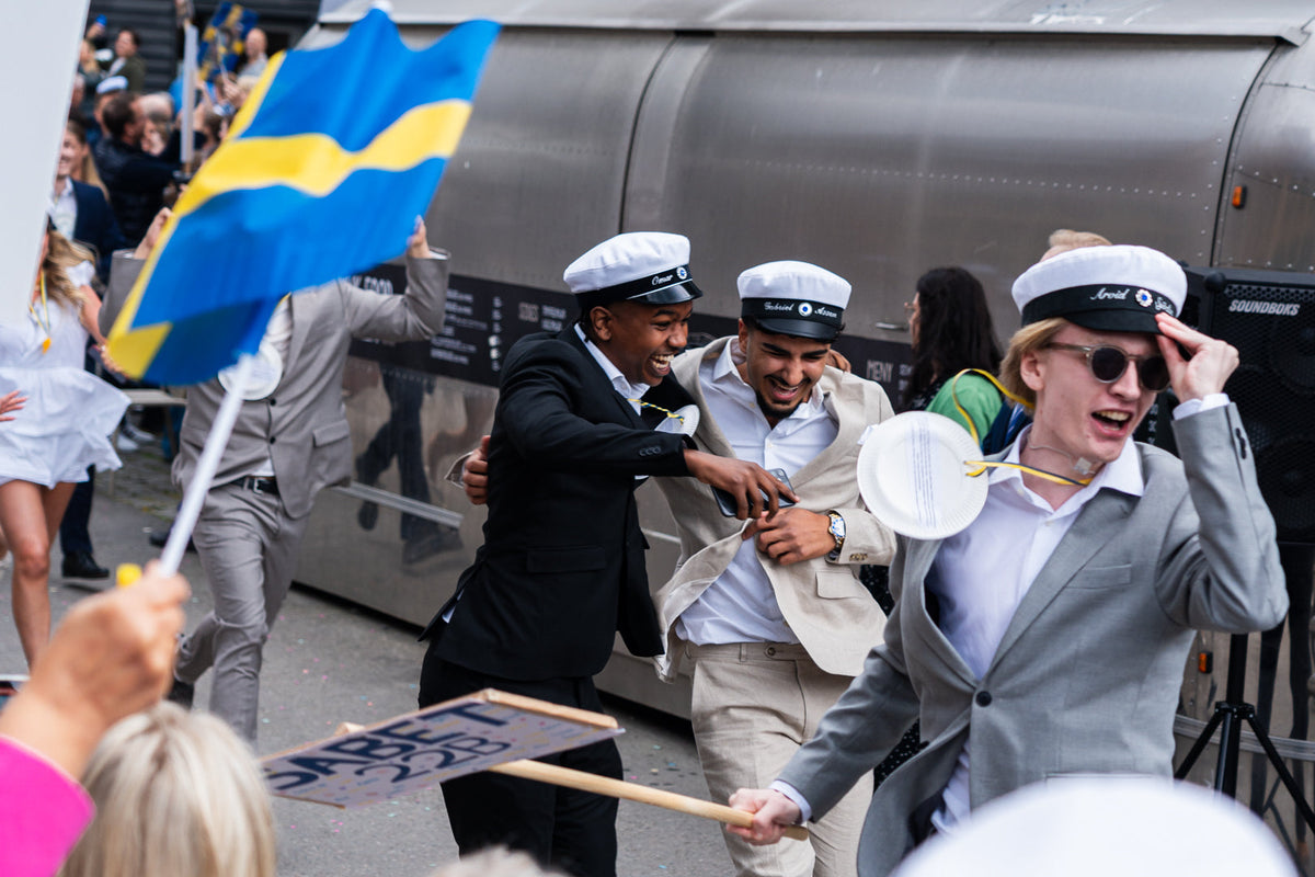 Studenter i vita studentmössor springer ut på studentdagen, en av dem med en svensk flagga