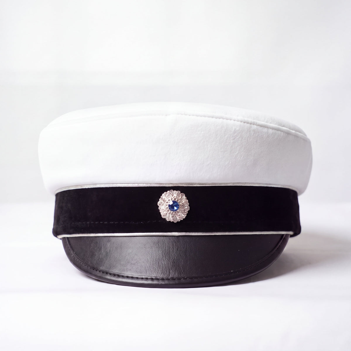 Studentmössa framifrån med vit kulle, svart sammetsband, svart skärm och kokard med blå sten i silverfattning