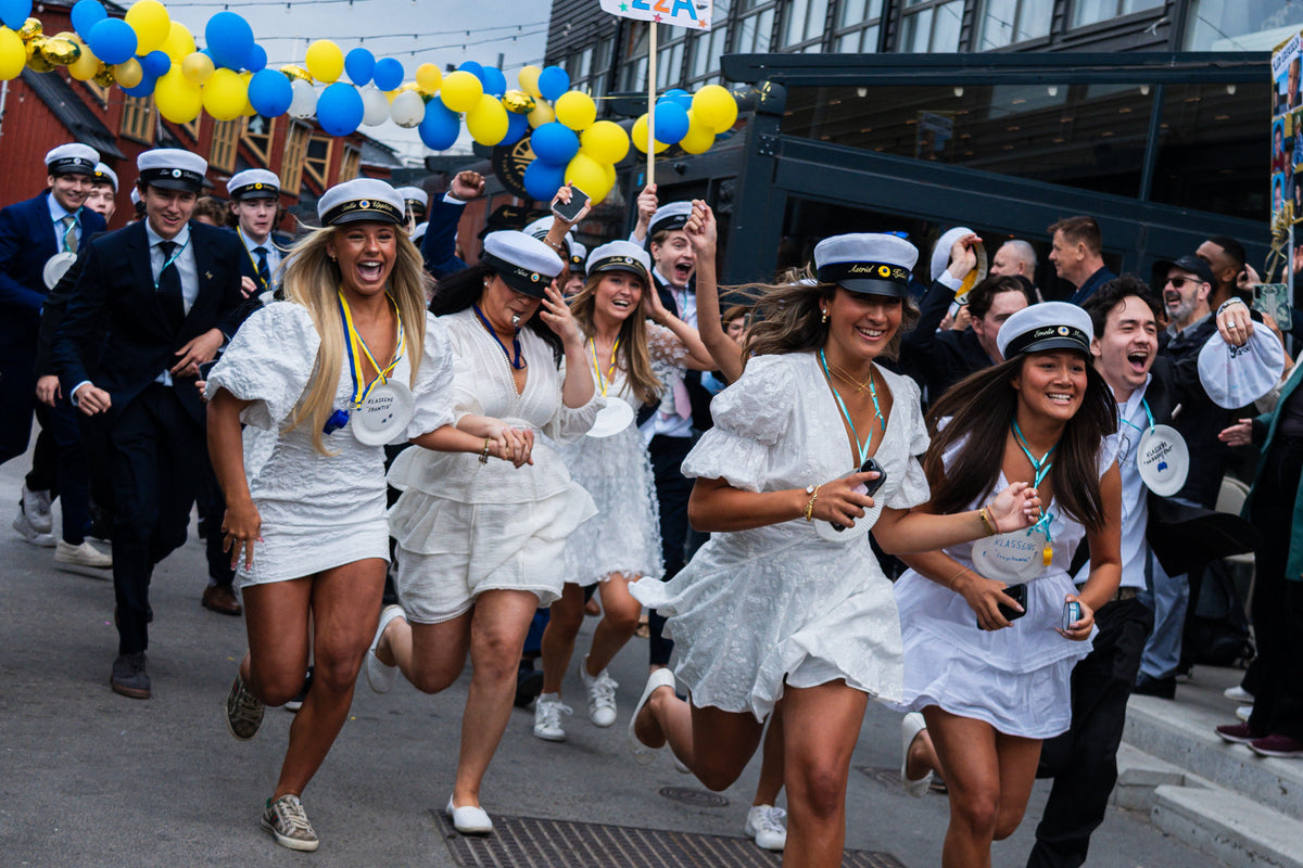 Studenter i vita studentmössor springer ut på studentdagen bland blågula ballonger