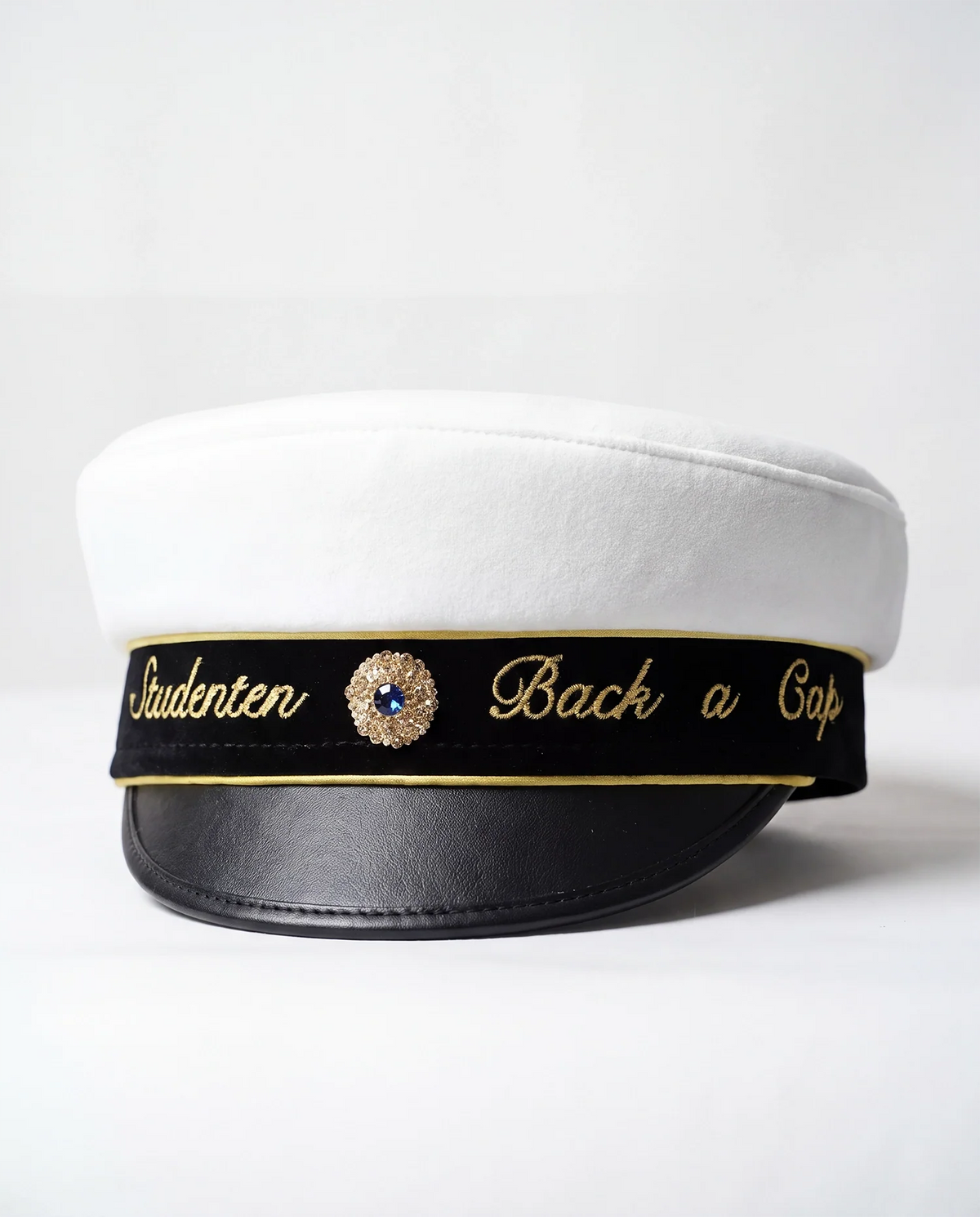 Text på studentmössan med ”Studenten” och ”Back a Cap” broderat i guld på var sida om kokarden